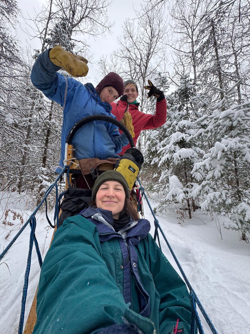 The image shows three people enjoying a winter outing, possibly dog sledding or riding on a sled. The person in the foreground is wearing a green jacket and hat, while the two people behind them are bundled up in winter gear. They are surrounded by snow-covered trees, suggesting a snowy, outdoor environment. The overall impression is one of winter fun and camaraderie.
